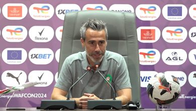 1000256458 | MCG24 | موقع إلكتروني مغربي شامل متجدد على مدار الساعة Man in a gray polo sits at a press conference table with two microphones, hands clasped, badge on chest, sponsor backdrop behind and a soccer ball to the side.