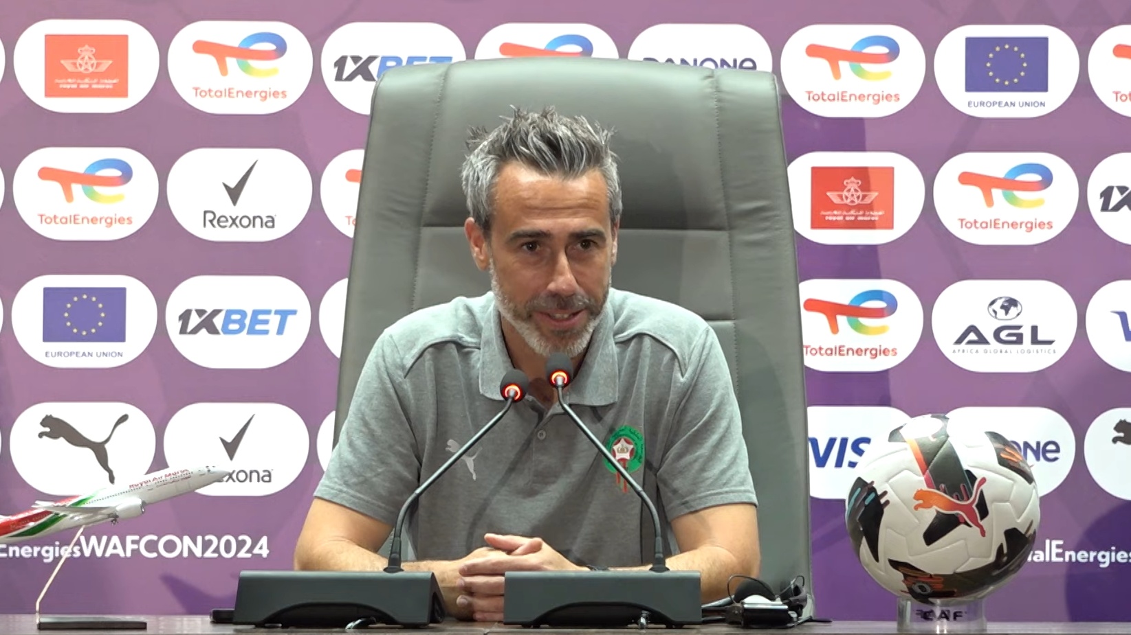 1000256458 | MCG24 | موقع إلكتروني مغربي شامل متجدد على مدار الساعة Man in a gray polo sits at a press conference table with two microphones, hands clasped, badge on chest, sponsor backdrop behind and a soccer ball to the side.