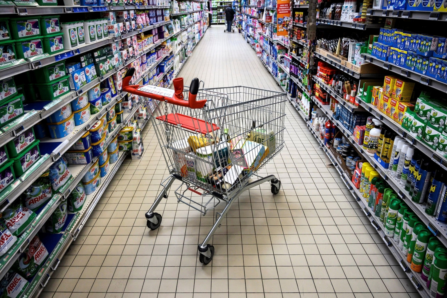 551545488 | MCG24 | موقع إلكتروني مغربي شامل متجدد على مدار الساعة Shopping cart filled with groceries in the center of a supermarket aisle lined with cleaning products on both sides.