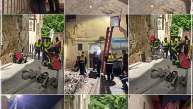 Firefighters in reflective gear repair a wall with a yellow ladder in a narrow street.