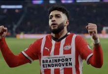 PSV Eindhoven player in red-and-white striped kit celebrates with fists raised on the pitch, Brainport Eindhoven sponsor on jersey.