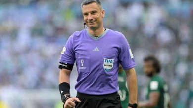 361 | MCG24 | موقع إلكتروني مغربي شامل متجدد على مدار الساعة Football referee in a purple kit with FIFA badge, headset mic, black armband, standing on a stadium pitch.