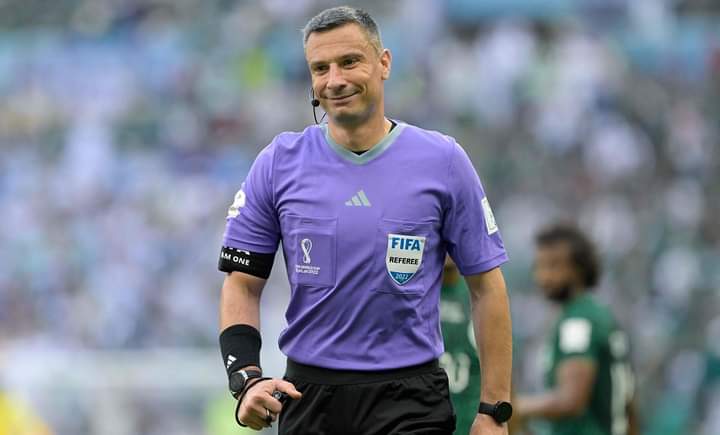 Football referee in a purple kit with FIFA badge, headset mic, black armband, standing on a stadium pitch.