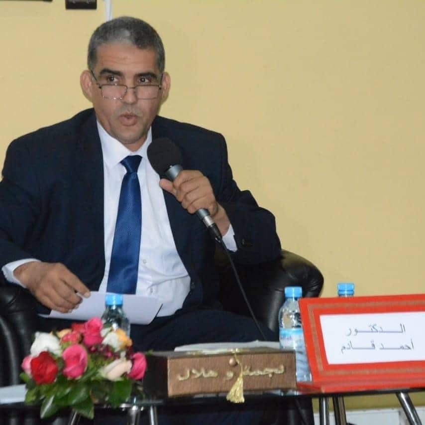 Man in a dark suit and blue tie sits in a panel chair, speaking into a handheld microphone at a formal event; bouquet of flowers and a name placard on the table in front of him (Arabic text visible).