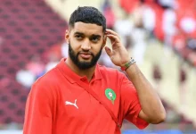 473938 | MCG24 | موقع إلكتروني مغربي شامل متجدد على مدار الساعة Bearded man in a red polo with a green Moroccan crest, touching his ear in a stadium.