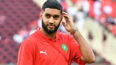 473938 | MCG24 | موقع إلكتروني مغربي شامل متجدد على مدار الساعة Bearded man in a red polo with a green Moroccan crest, touching his ear in a stadium.