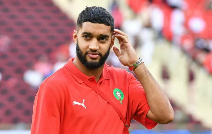 Bearded man in a red polo with a green Moroccan crest, touching his ear in a stadium.