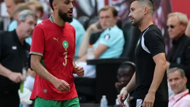 Two male athletes converse on a sports court; one in a red jersey with number 3 and green shorts, the other in a black kit.