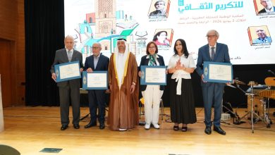 Group of six people on a stage holding blue-framed certificates at a formal ceremony, backdrop shows Arabic text and illustrations of buildings.