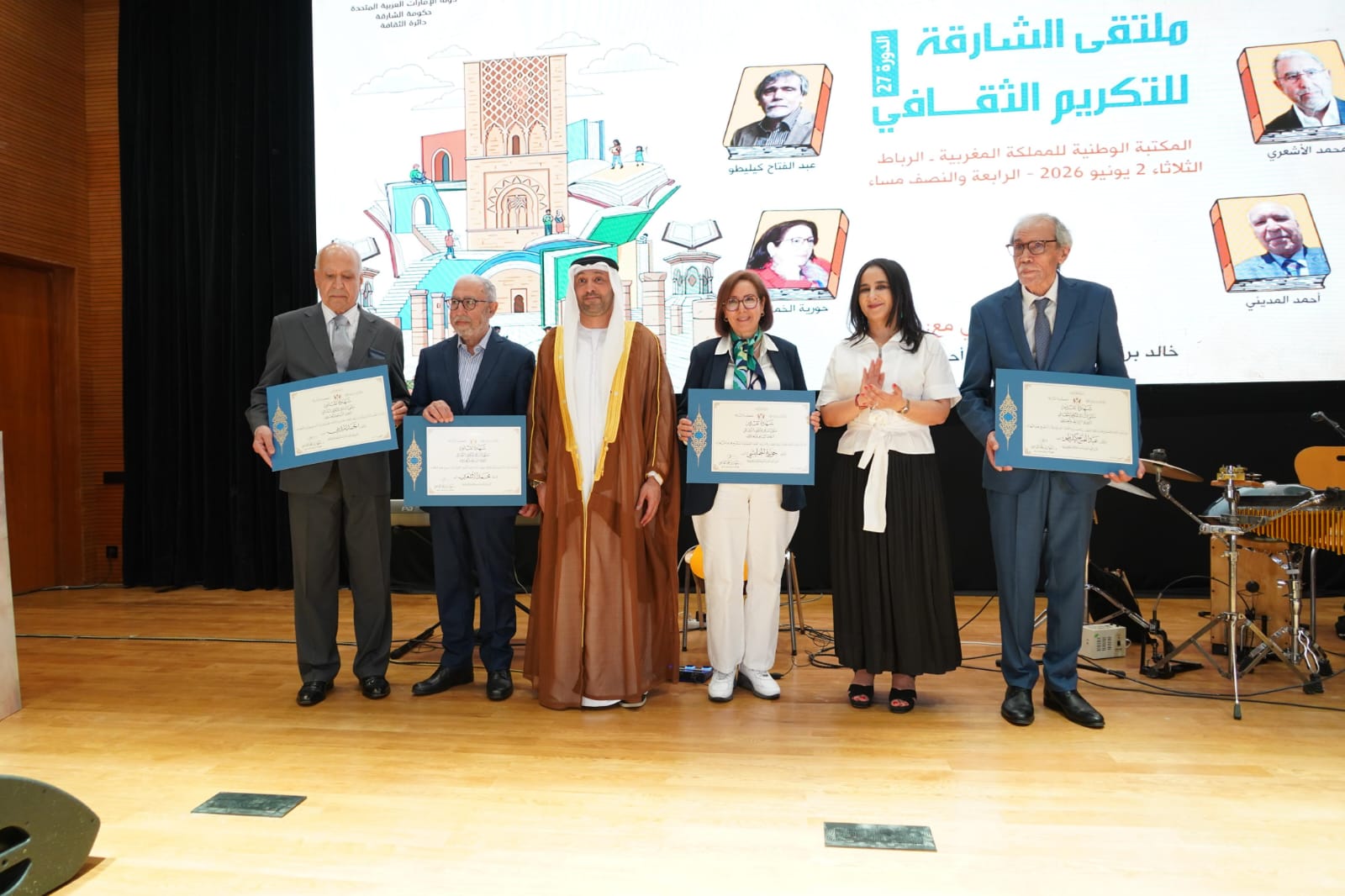 Group of six people on a stage holding blue-framed certificates at a formal ceremony, backdrop shows Arabic text and illustrations of buildings.
