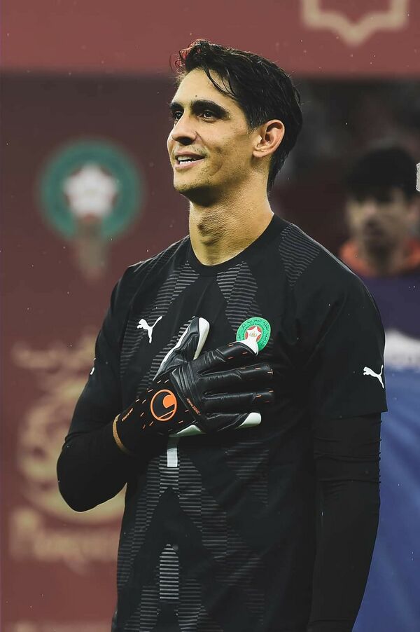 Soccer goalkeeper in a black jersey with gloves, hand on chest, smiling on the field.