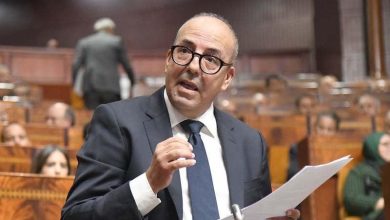 Man in a dark suit and glasses speaks into a microphone while holding a document in a wooden chamber setting.