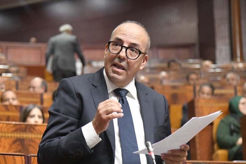 Man in a dark suit and glasses speaks into a microphone while holding a document in a wooden chamber setting.