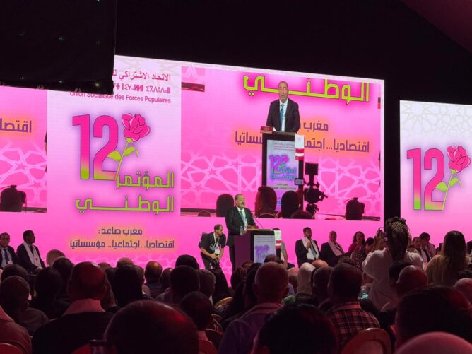 68f271226978e-668x501 | MCG24 | موقع إلكتروني مغربي شامل متجدد على مدار الساعة Speaker at a podium on a bright pink stage with Arabic text and a large '12' rose logo; crowded audience watching.
