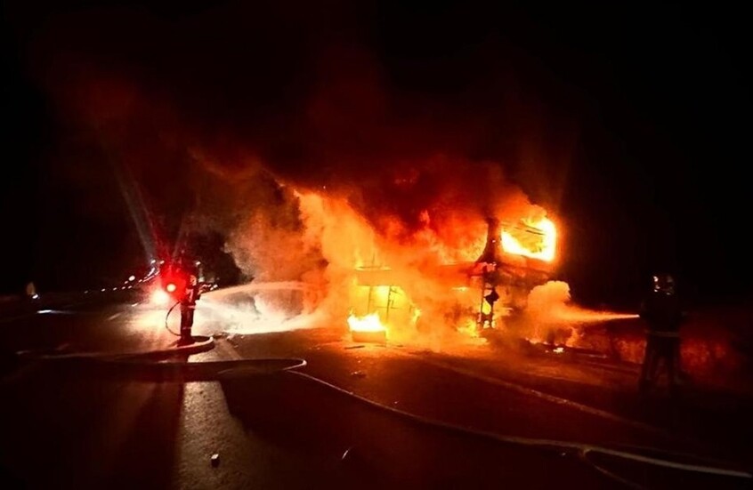 Night-time scene of a burning building with flames engulfing the structure as firefighters spray water from hoses nearby.