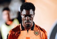 Portrait of a footballer in an orange jersey with a club crest, looking downward in concentration, head bowed.