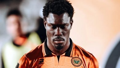 Portrait of a footballer in an orange jersey with a club crest, looking downward in concentration, head bowed.