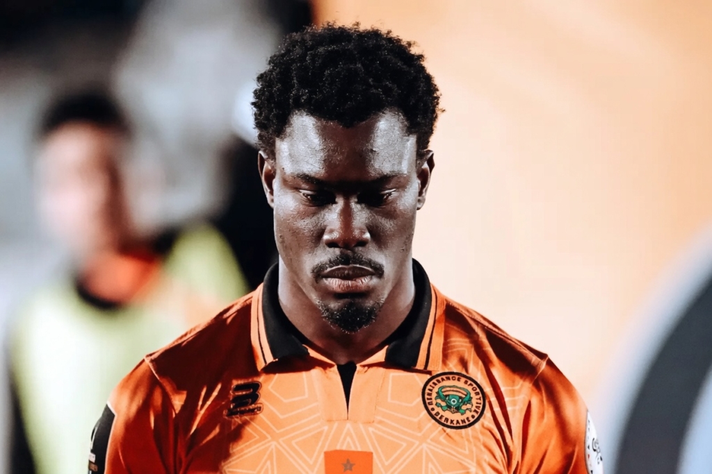 Portrait of a footballer in an orange jersey with a club crest, looking downward in concentration, head bowed.