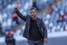 Man in a dark blazer and black shirt waves while walking across a stadium field, with blurred empty seats in the background onlookers far away.