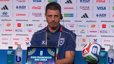 Coach at a FIFA World Cup press conference, seated with a laptop displaying 'San Francisco Bay Area' and a soccer ball on the table, backdrop full of sponsor logos.