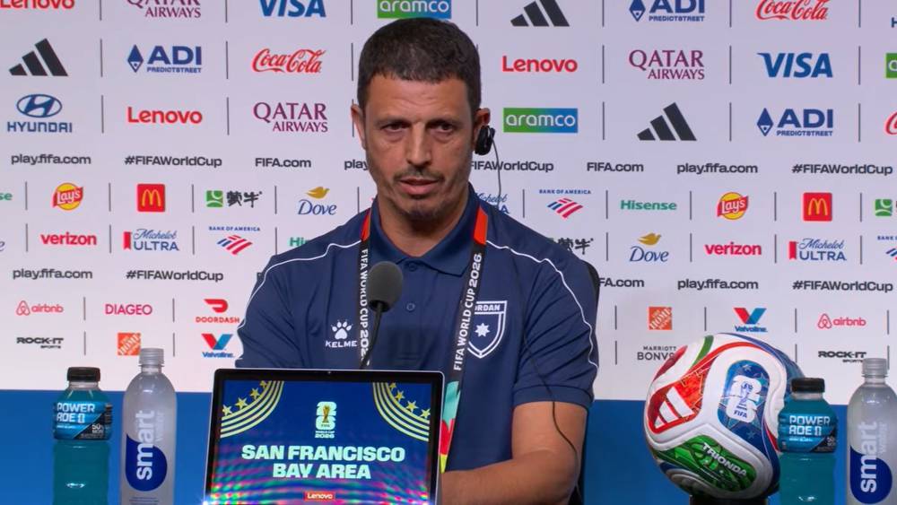 6a326fd8fe191bd3077e9030_default | MCG24 | موقع إلكتروني مغربي شامل متجدد على مدار الساعة Coach at a FIFA World Cup press conference, seated with a laptop displaying 'San Francisco Bay Area' and a soccer ball on the table, backdrop full of sponsor logos.