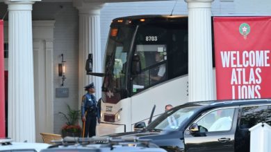 White coach bus numbered 8703 parked at a columned entrance; a uniformed security officer stands nearby, next to a red banner that reads 'Welcome Atlas Lions'.
