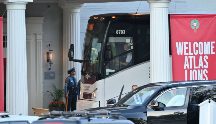 White coach bus numbered 8703 parked at a columned entrance; a uniformed security officer stands nearby, next to a red banner that reads 'Welcome Atlas Lions'.