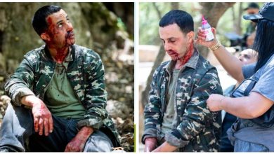 Man in camouflage jacket sits outdoors with blood on his face and hands (prop injuries).