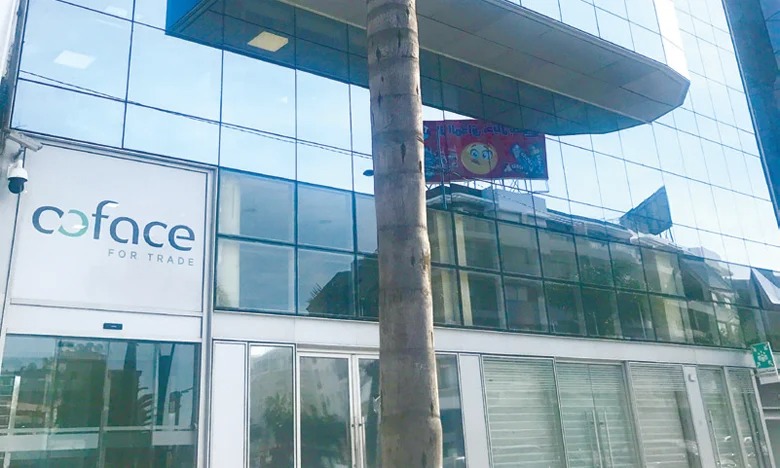Glass-front office building with a Coface for Trade sign on the left and a palm tree in the foreground. There’s a banner with a smiley face overhead.