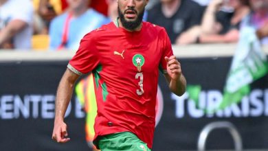 79F4F799-AFC6-4037-88BD-C922B66B5C90 | MCG24 | موقع إلكتروني مغربي شامل متجدد على مدار الساعة Soccer player in a red jersey with number 3 and green shorts running on the pitch during a match, crowd in the stands behind him.