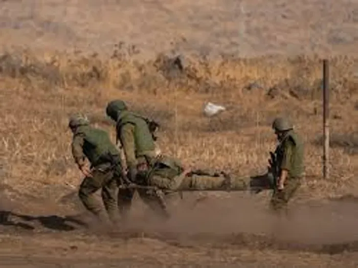 Four soldiers in green uniforms carry a wounded person on a stretcher across a dusty field at a barren outdoor area.