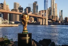 FIFA World Cup Trophy on a pedestal beside the river with the Manhattan skyline and Brooklyn Bridge behind.