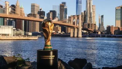 FIFA World Cup Trophy on a pedestal beside the river with the Manhattan skyline and Brooklyn Bridge behind.