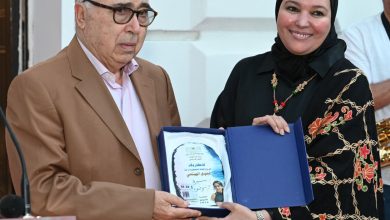 Older man in a brown suit and glasses receives an award from a smiling woman in a black hijab; the blue case opens to reveal the plaque inside.