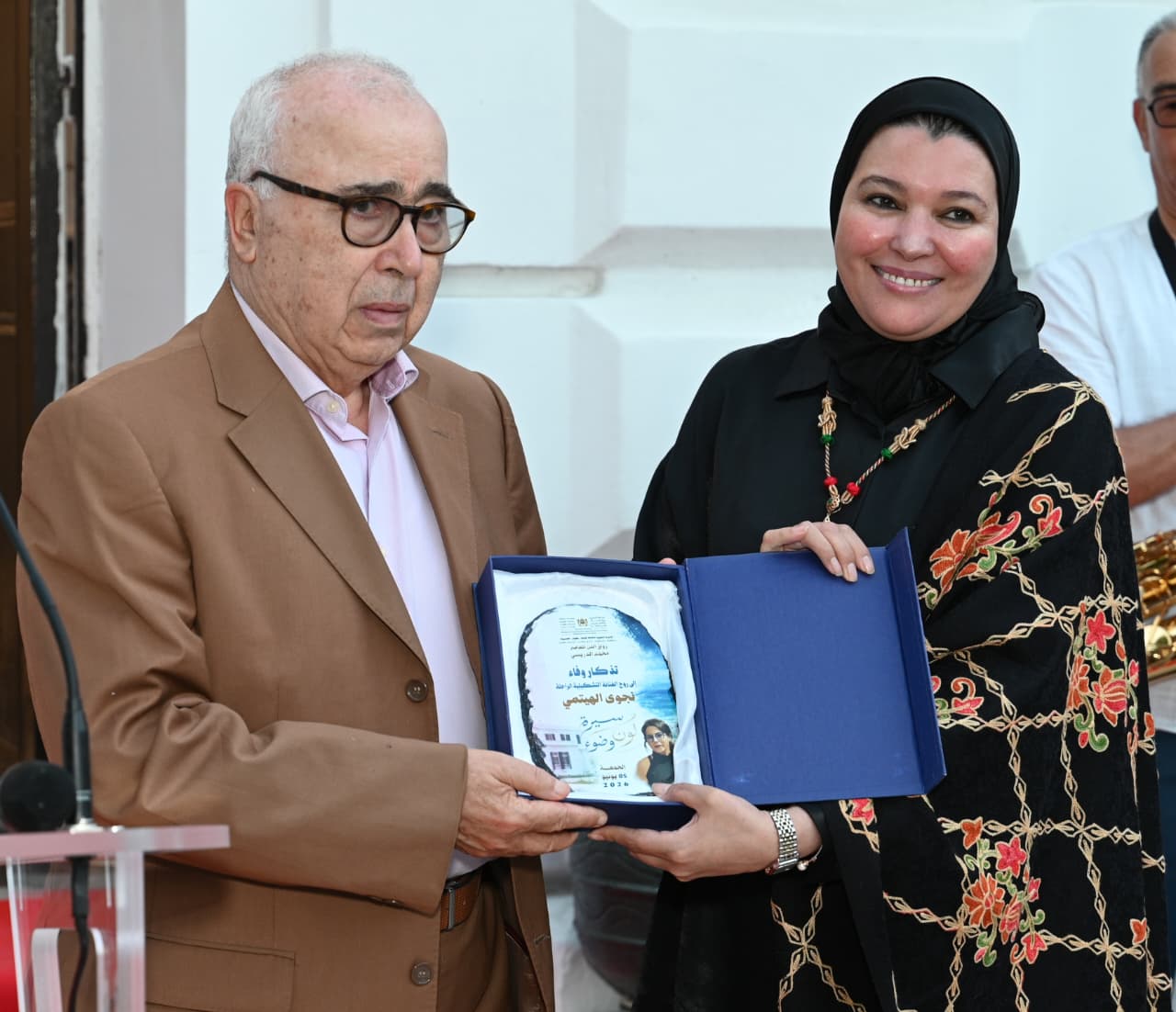 8a549361-ddde-4f21-8ddc-7ca53cf06bb1 | MCG24 | موقع إلكتروني مغربي شامل متجدد على مدار الساعة Older man in a brown suit and glasses receives an award from a smiling woman in a black hijab; the blue case opens to reveal the plaque inside.