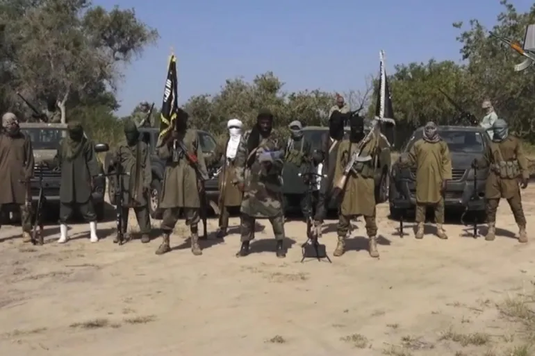 Armed militants in desert, lined up with rifles, wearing masks, with black flags and cars in the background.
