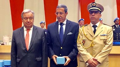 Three men in formal attire at a ceremony; center man holds a light blue box, flanked by a suited man and a uniformed officer.