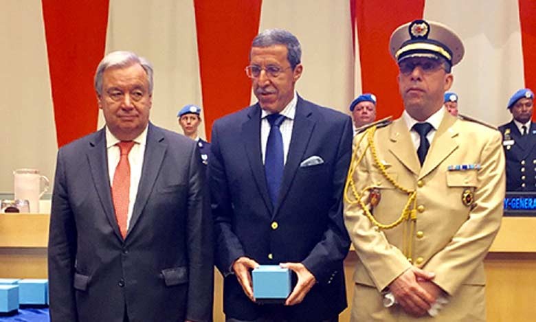 Three men in formal attire at a ceremony; center man holds a light blue box, flanked by a suited man and a uniformed officer.