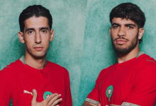 Two men in red jerseys with green badges, standing against a green textured background, posing confidently side by side.