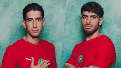 Two men in red jerseys with green badges, standing against a green textured background, posing confidently side by side.