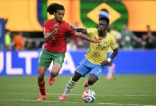 bouaddi | MCG24 | موقع إلكتروني مغربي شامل متجدد على مدار الساعة Two soccer players—one in yellow Brazil kit and one in red—jostle for the ball on a green field as a Brazilian flag stands in the background.