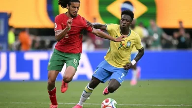bouaddi | MCG24 | موقع إلكتروني مغربي شامل متجدد على مدار الساعة Two soccer players—one in yellow Brazil kit and one in red—jostle for the ball on a green field as a Brazilian flag stands in the background.