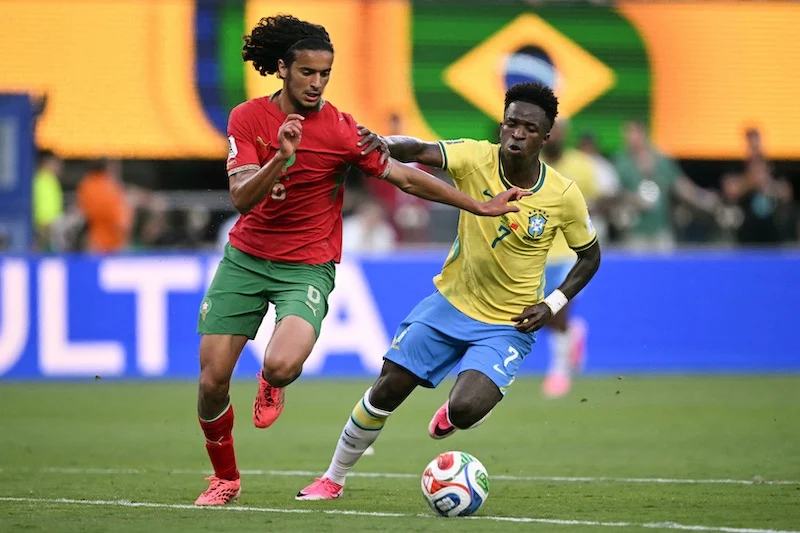 bouaddi | MCG24 | موقع إلكتروني مغربي شامل متجدد على مدار الساعة Two soccer players—one in yellow Brazil kit and one in red—jostle for the ball on a green field as a Brazilian flag stands in the background.