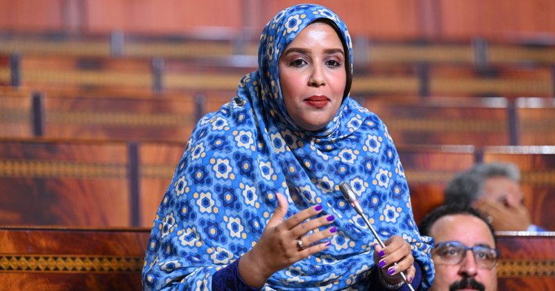 BAH_8932-scaled | MCG24 | موقع إلكتروني مغربي شامل متجدد على مدار الساعة Woman wearing a blue patterned hijab speaks into a microphone in a wood-paneled legislative chamber.