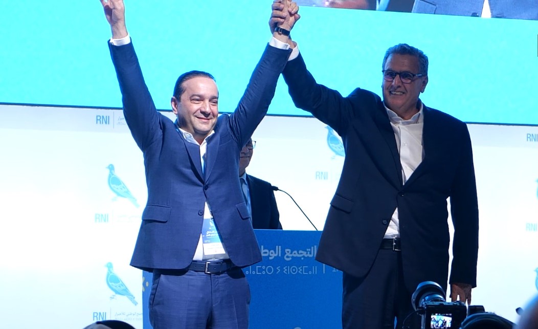 Two men in dark suits celebrate on stage, lifting their clasped hands in victory.