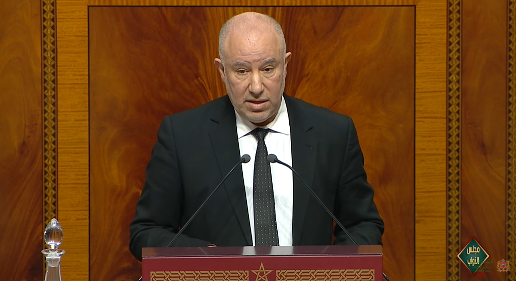 A man in a black suit and tie speaks from a wooden podium with two microphones in front of him.