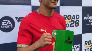 diaz | MCG24 | موقع إلكتروني مغربي شامل متجدد على مدار الساعة Man in a red soccer jersey holds a green trophy with 'FC26' branding at a 'The World's Game' event backdrop, smiling for the camera in a prize win moment.
