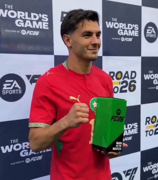 diaz | MCG24 | موقع إلكتروني مغربي شامل متجدد على مدار الساعة Man in a red soccer jersey holds a green trophy with 'FC26' branding at a 'The World's Game' event backdrop, smiling for the camera in a prize win moment.