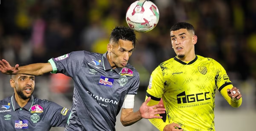 Two soccer players from opposing teams jump for a header as the ball flies overhead, one in a gray kit on the left and the other in a bright yellow kit on the right.