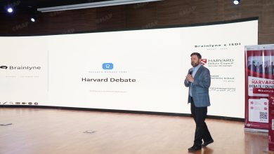 Male presenter in a blue checkered suit speaks into a microphone on a stage with a large curved screen showing 'Harvard Debate' and Brainlyne branding.